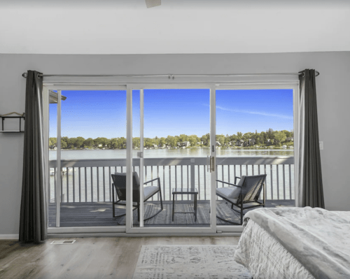 A serene view of a lake from a bedroom, featuring sliding glass doors and two chairs on a balcony.