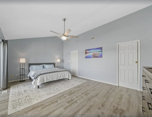 Spacious bedroom with a gray bed, ceiling fan, and light wood flooring, featuring a large window and decorative artwork.