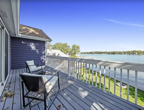 A serene lakeside balcony with two chairs, overlooking calm water and green grass under a clear blue sky.