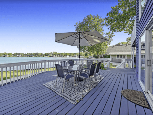 A lakeside deck with a table, chairs, and an umbrella, overlooking calm water and trees under a clear blue sky.