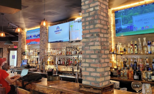 A cozy bar with a brick wall, shelves of liquor, and multiple screens showing sports events.
