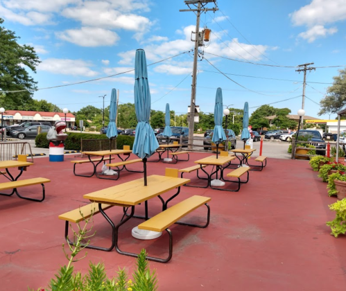 Outdoor dining area with wooden picnic tables and blue umbrellas under a clear sky, surrounded by greenery.