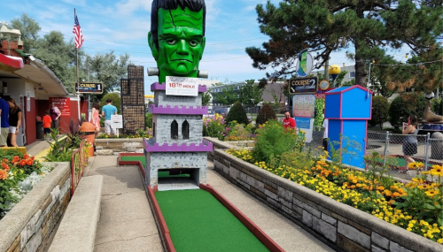 A mini-golf course featuring a large green Frankenstein head atop a castle structure, surrounded by colorful flowers.