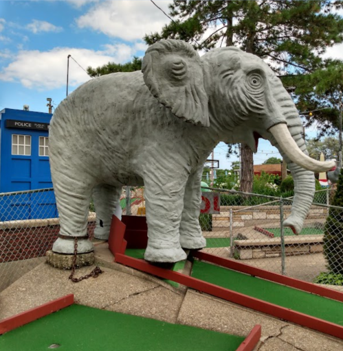 A large concrete elephant sculpture stands on a mini-golf course, surrounded by green turf and trees.