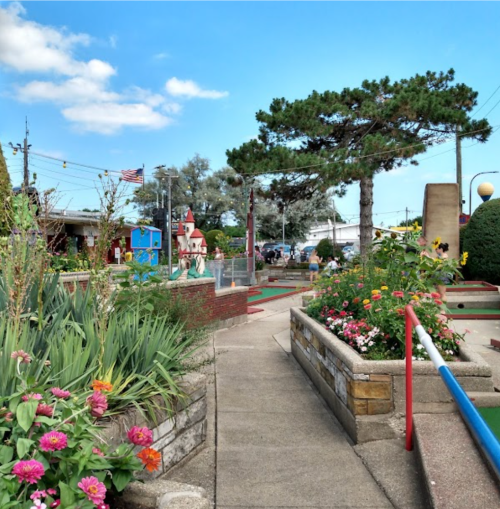 A colorful mini-golf course with flowers, trees, and a clear blue sky in the background.