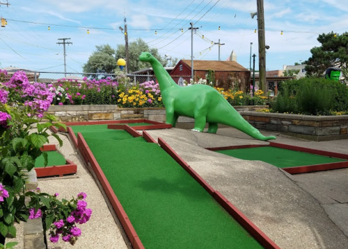 A green dinosaur statue stands on a mini-golf course surrounded by colorful flowers and greenery.