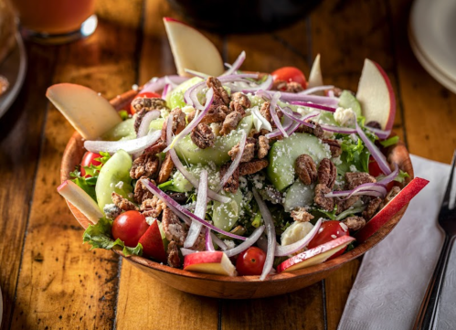 A fresh salad in a wooden bowl, featuring greens, cucumbers, tomatoes, apples, red onions, and nuts.
