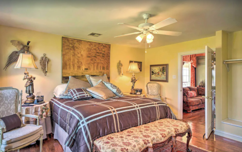 Cozy bedroom with a large bed, decorative lamps, artwork, and a view of an adjoining room. Warm yellow walls.