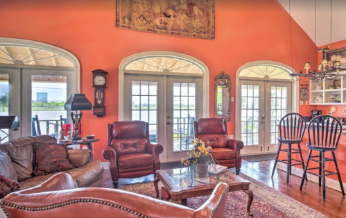 Cozy living room with orange walls, leather chairs, a glass table, and large windows overlooking a patio.