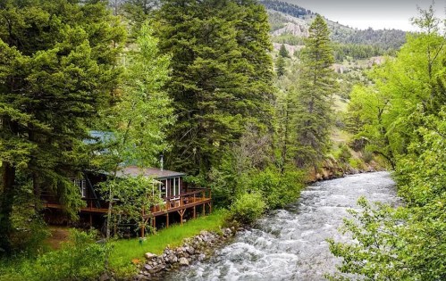 A cozy cabin by a rushing river, surrounded by lush green trees and mountains in the background.