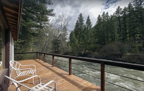 A wooden deck with white chairs overlooks a rushing river surrounded by lush green trees under a cloudy sky.