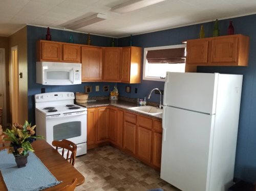 A cozy kitchen with wooden cabinets, white appliances, and a dining table, featuring colorful decor on the shelves.