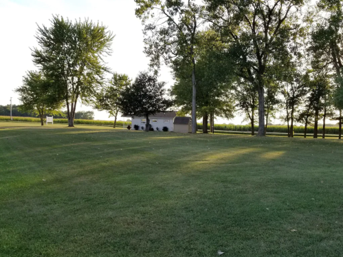 A grassy area with trees and a small building in the background, surrounded by fields under a clear sky.