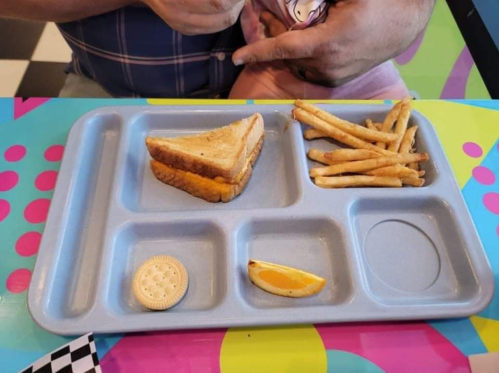 A blue cafeteria tray with a grilled cheese sandwich, fries, a cracker, and a slice of orange on a colorful table.
