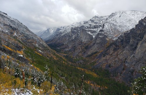 A scenic mountain valley with snow-capped peaks, autumn foliage, and a cloudy sky.