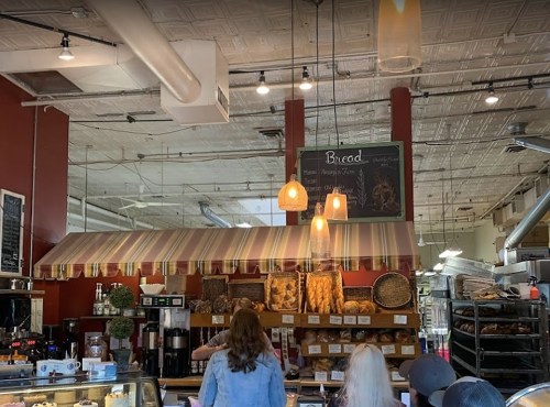 A cozy bakery interior with a striped awning, shelves of bread, and warm lighting. Customers are browsing the selection.