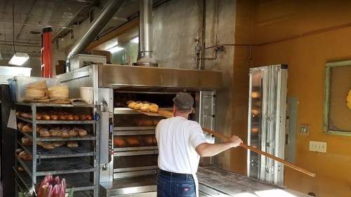 A baker using a long paddle to place bread into a large oven in a bakery. Shelves of baked goods are visible nearby.