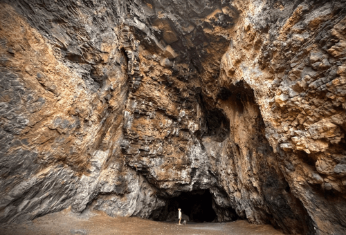 A person stands at the entrance of a large, rocky cave with textured walls and a dark interior.