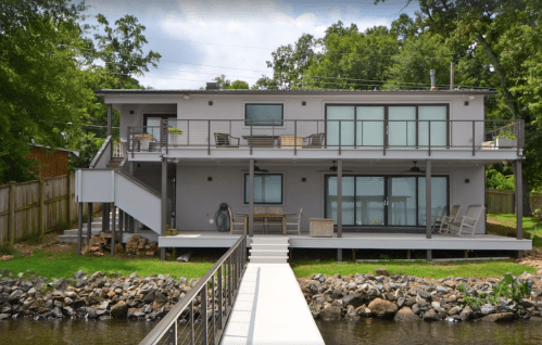Modern two-story house by a lake, featuring large windows, a deck, and a wooden walkway leading to the water.