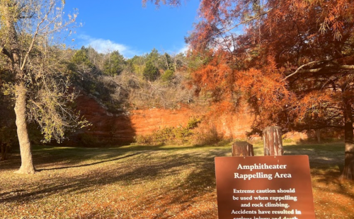 Sign for the Amphitheater Rappelling Area, surrounded by trees and a red rock cliff in the background.