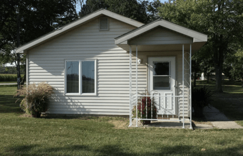 A small, light-colored house with a front porch and large windows, surrounded by grass and trees.