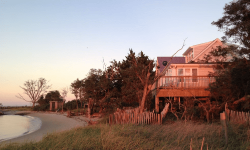 A beach house at sunset, surrounded by trees and tall grass, with a calm shoreline in the foreground.