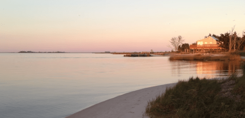 A serene waterfront scene at sunset, featuring a house, calm water, and gentle shoreline with grass.