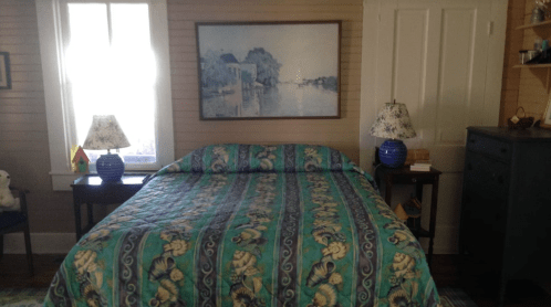 A cozy bedroom featuring a floral quilted bedspread, two lamps, and a large painting above the bed.