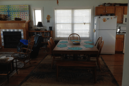 A cozy dining area with a wooden table, chairs, and a view of a kitchen and living space. Sunlight filters through blinds.