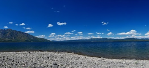 A panoramic view of a clear blue lake surrounded by mountains and a rocky shoreline under a bright sky with fluffy clouds.