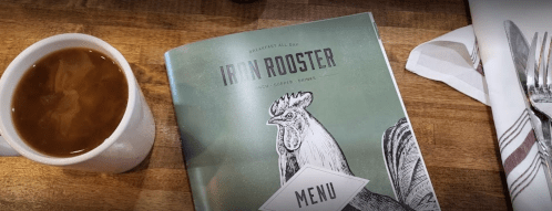 A coffee cup beside an open menu featuring a rooster illustration on a wooden table.