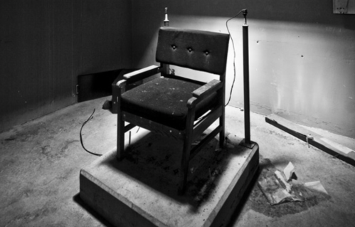 A dimly lit, empty electric chair in a stark, abandoned room with scattered debris on the floor.