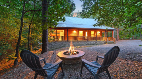 A cozy cabin surrounded by trees, featuring a fire pit and two chairs in front, illuminated at dusk.