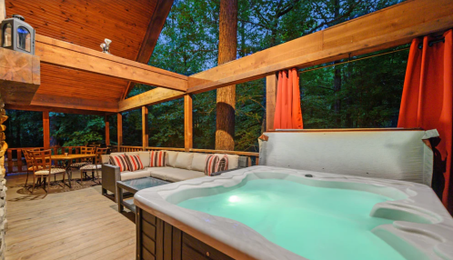 Cozy outdoor space featuring a hot tub, seating area, and dining table surrounded by trees at dusk.