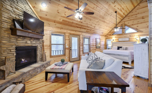 Cozy cabin interior with wooden walls, a stone fireplace, a comfortable sofa, and a large bed under a vaulted ceiling.