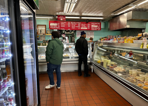 Two customers browse a deli with glass display cases filled with various food items and a menu on the wall.
