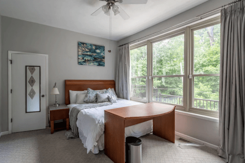 Bright bedroom with a large window, a bed with decorative pillows, a wooden desk, and a nightstand.