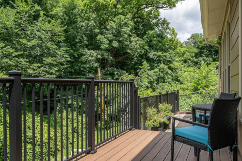 A wooden deck with black railings overlooks a lush green forest under a cloudy sky. A chair is visible on the deck.