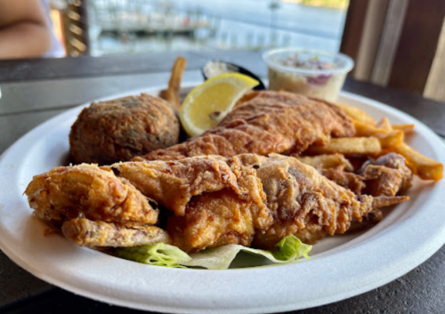 A plate of fried fish, fries, a hushpuppy, and a lemon wedge, served with a side of coleslaw.