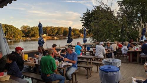 A lively outdoor dining area by a river, with people enjoying meals at picnic tables under blue umbrellas.