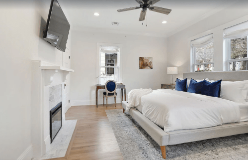 Bright, modern bedroom with a large bed, TV, desk, and fireplace, featuring white walls and blue accents.