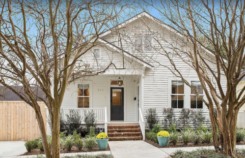 A charming white house with a front porch, surrounded by trees and colorful flower pots, set in a landscaped yard.