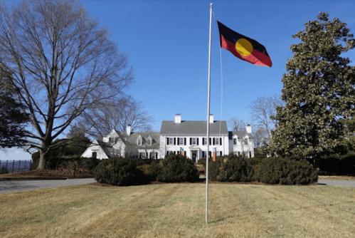 A large white house with a flag featuring a circle and colors, surrounded by green bushes and a clear blue sky.