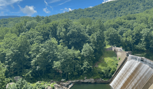 A scenic view of a dam cascading water into a lush green forested area under a blue sky with scattered clouds.