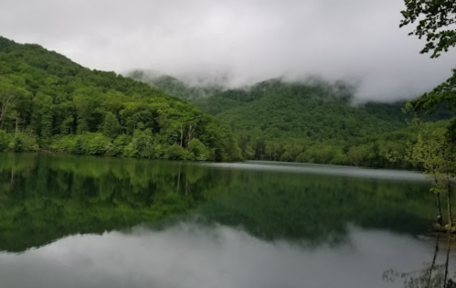 A serene lake surrounded by lush green mountains under a cloudy sky, with reflections on the water's surface.
