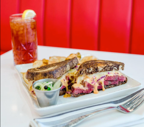 A plate with a grilled sandwich, chips, and a drink, set against a bright red background.