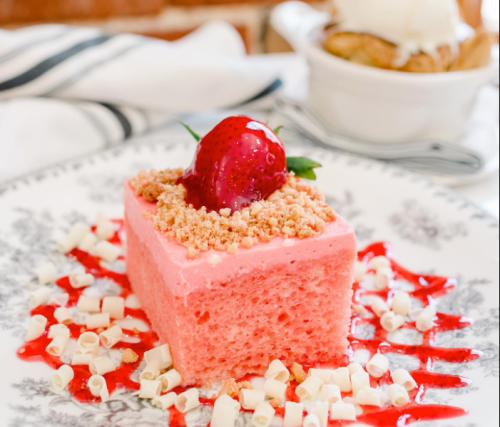 A slice of pink dessert topped with a strawberry and crumbs, drizzled with red sauce and white chocolate shavings.