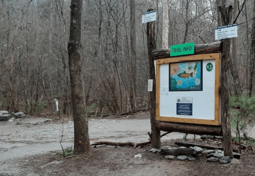 Trail information sign at a wooded area, featuring a map and details, surrounded by bare trees and a gravel path.