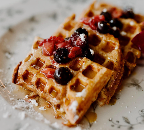 Two waffles topped with mixed berries and powdered sugar, drizzled with syrup on a decorative plate.