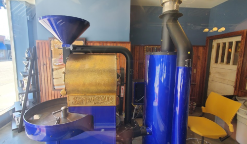 A vintage coffee roaster in a cozy café setting, featuring a blue and gold design with a funnel and exhaust pipe.